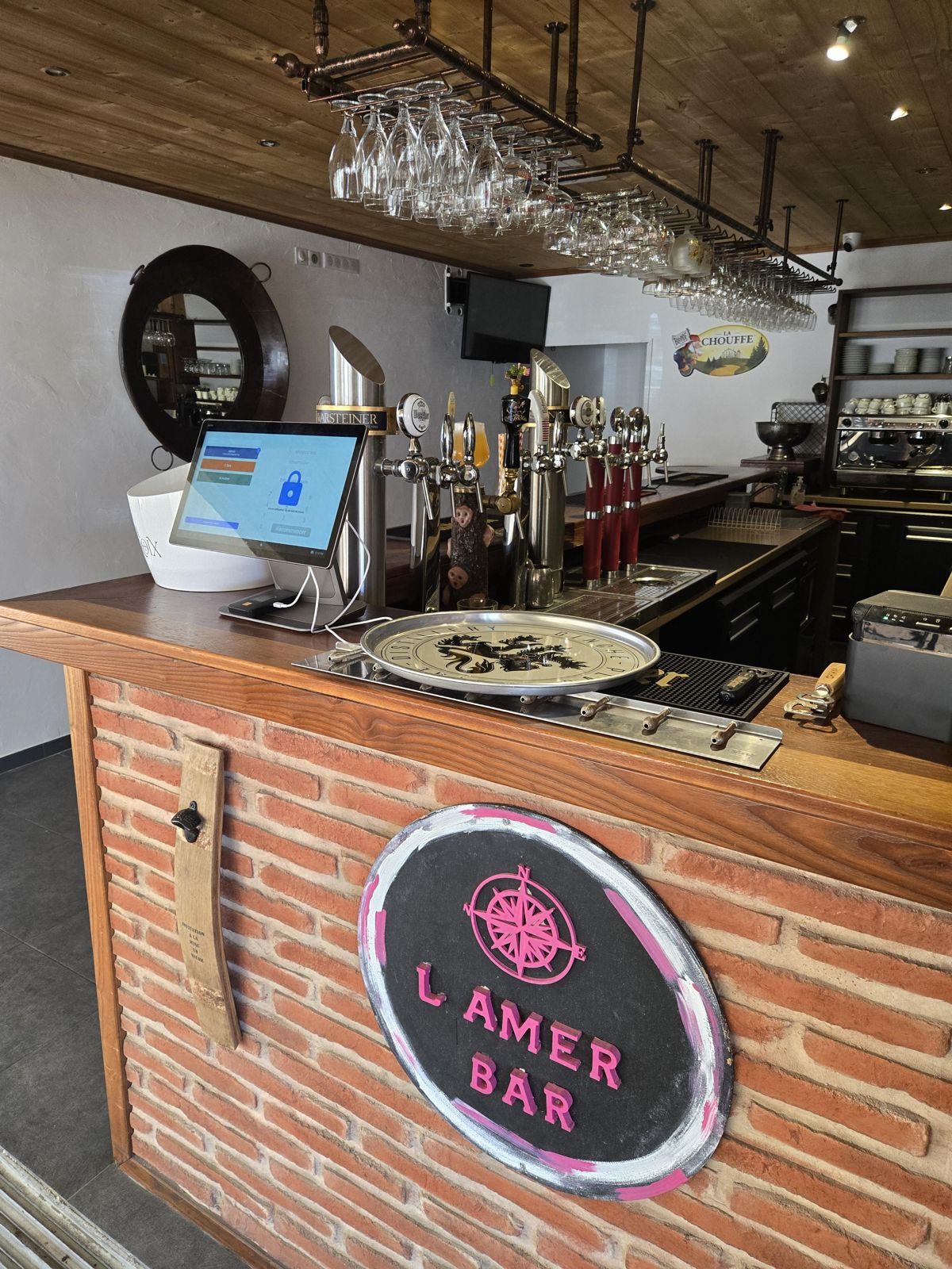 Le comptoir en briques, ses tireuses à bière, ses verres suspendus et l'enseigne ronde rose « L'Amer Bar »
