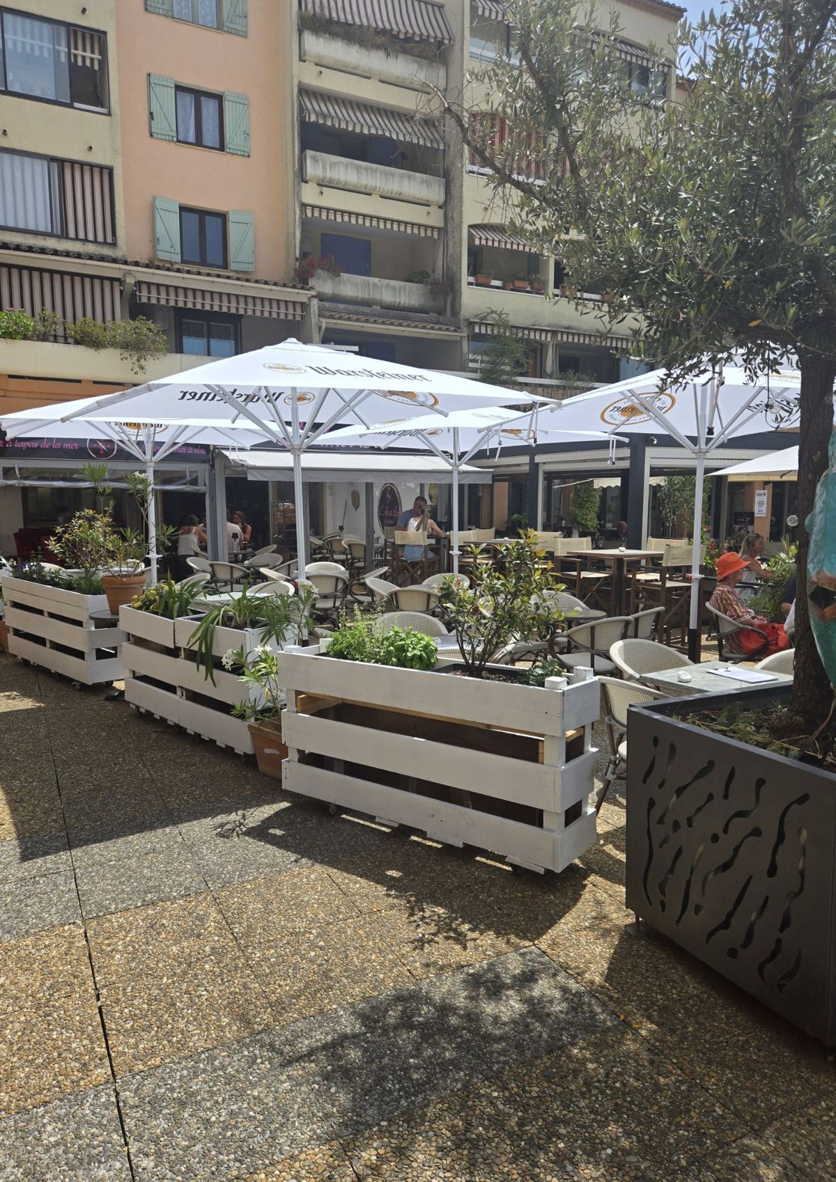 La terrasse sous de grands parasols, jardinières en palettes blanches et olivier, au pied des immeubles du village